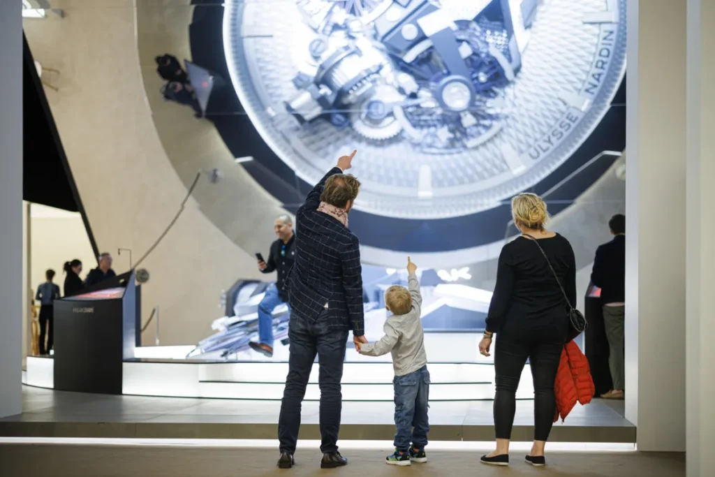Watches and Wonders 2026, visiteurs découvrant une installation horlogère géante au salon de Genève, moment en famille devant une œuvre mécanique Ulysse Nardin.