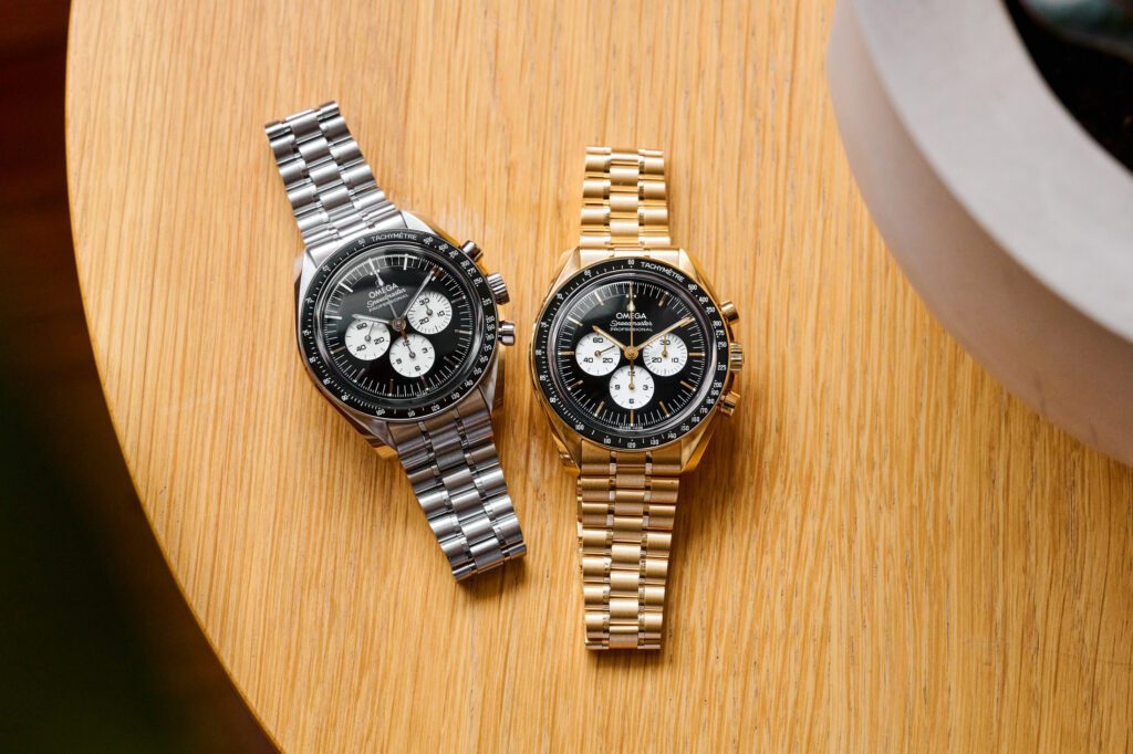 nouvelle omega speedmaster, montre Omega Reverse Panda posée sur une table en bois, modèle Moonwatch en acier avec cadran noir et sous-compteurs blancs, à côté de la version en or jaune.